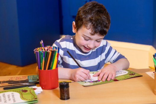 young boy sat drawing art a table with a red pot filled with colouring pencils next to him
