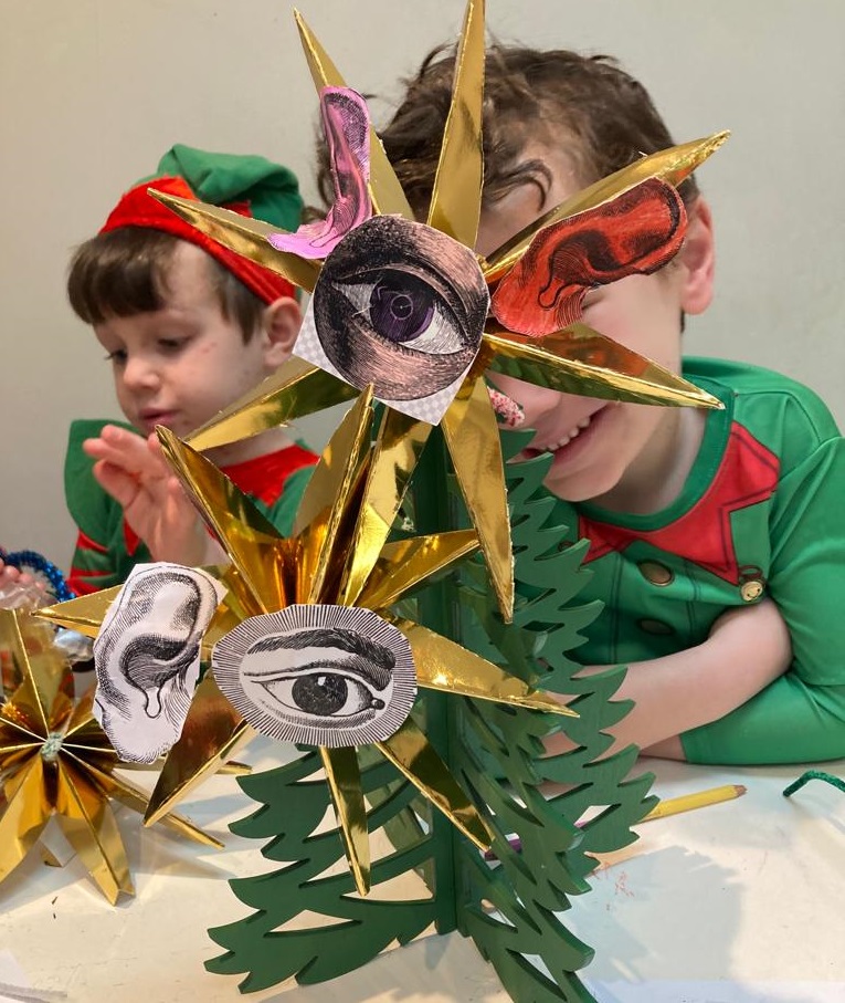 Two children dressed in elf outfits are sitting beside a craft Christmas tree with Surreal decorations on it
